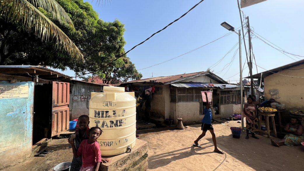 A community scene in Freetown, Sierra Leone showing children near a water tank, with a newly installed solar-powered LED streetlight visible on a pole among modest buildings.