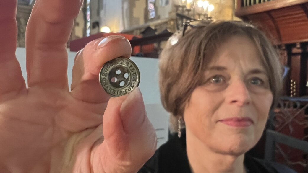 A woman holds up a small metal button with four holes and text around the rim, photographed inside what appears to be a church or historic building.