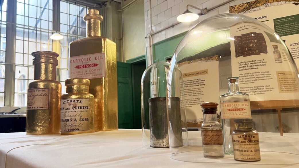 Museum display showing gold-foil wrapped chocolate replica medicine bottles alongside original glass bottles from Tyntesfield's medical cabinet, displayed under glass domes with explanatory text panels.
