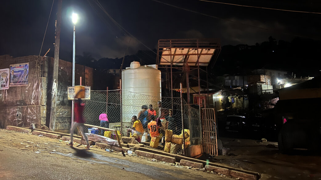 Nighttime street scene in Freetown showing a solar-powered LED streetlight illuminating a water collection point where people gather near a large white water tank.