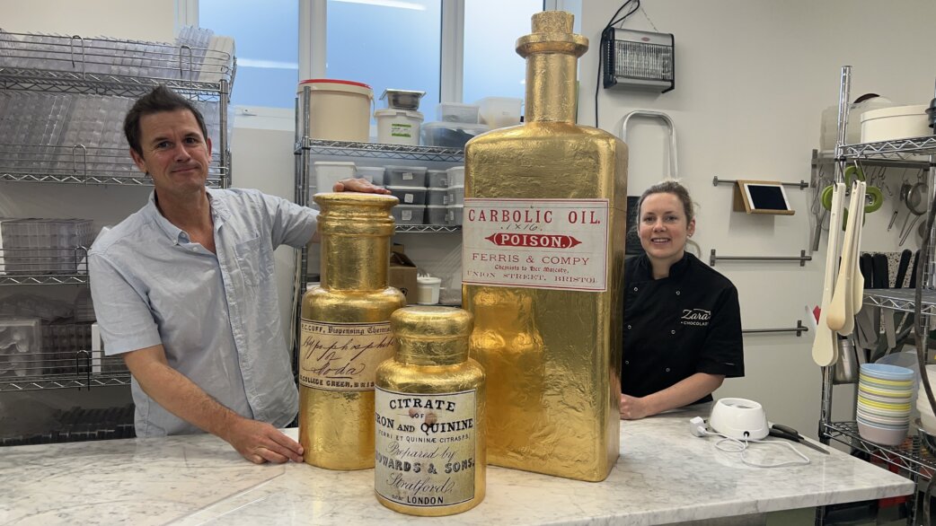 Two people stand in a commercial kitchen beside three gold foil-wrapped chocolate bottle replicas, the largest approximately one metre tall, modelled on Victorian medicine bottles.
