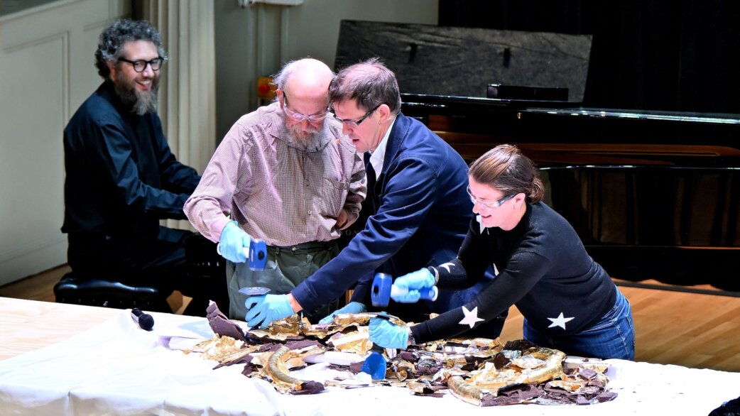 Four people wearing blue gloves use hammers and chisels to break apart gold foil-wrapped chocolate sculptures on a white-covered table, with a grand piano visible behind them.