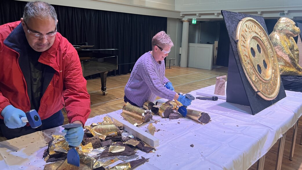 Two people wearing blue gloves use hammers and chisels to break apart gold foil-wrapped chocolate sculptures on a white-clothed table, with a gilded chocolate ship's wheel visible nearby.