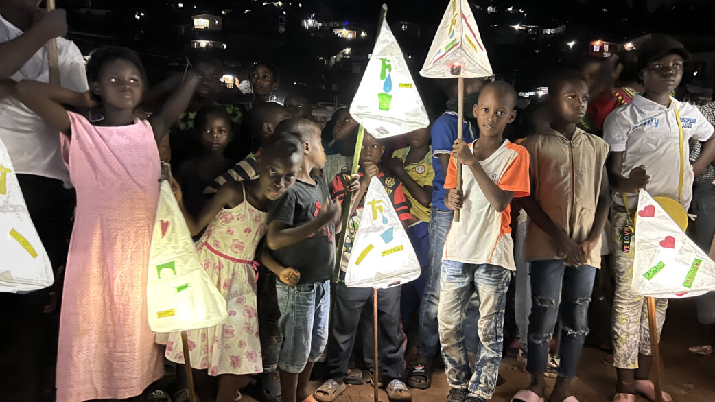 Children in Freetown, Sierra Leone at night holding illuminated triangular paper lanterns decorated with colourful drawings, part of the Power of Light community project and parade.