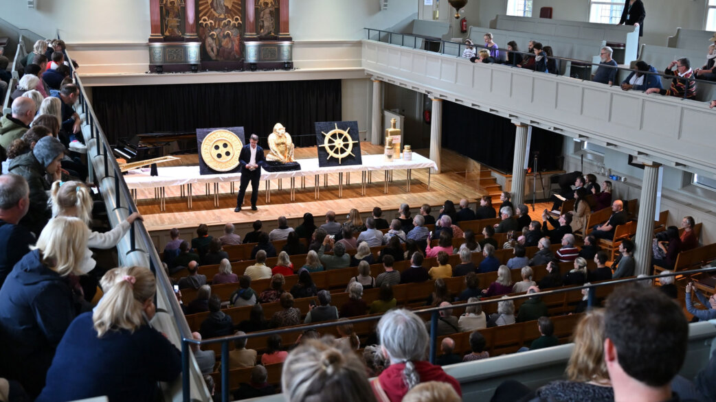 An audience seated in a church hall watches a presenter on stage beside large gold-foiled chocolate sculptures including a ship's wheel, button, and seated figure during the Edible Histories event.