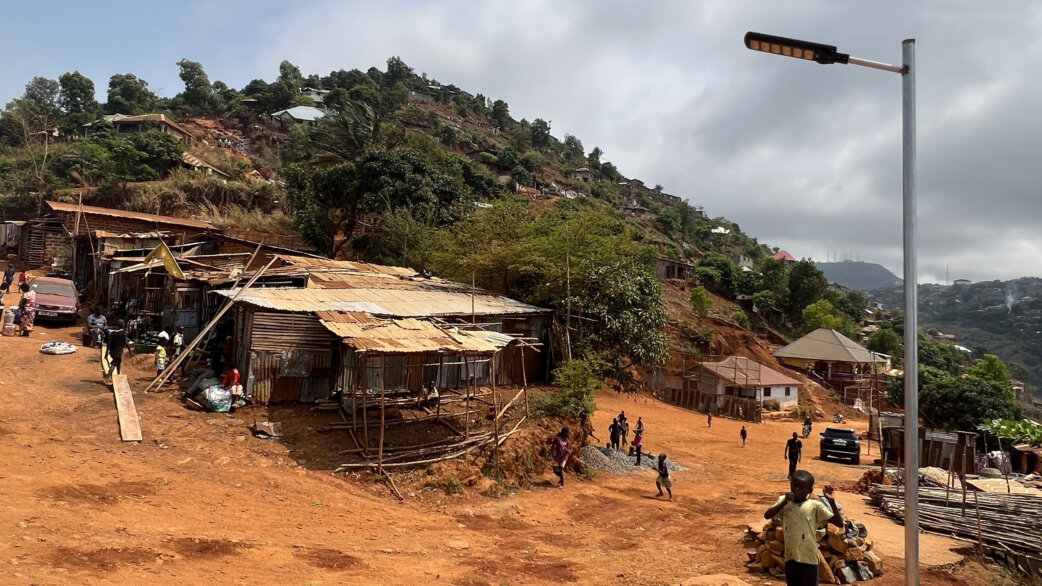 A hillside informal settlement in Freetown, Sierra Leone, with corrugated metal structures on red earth and a newly installed solar-powered LED streetlight in the foreground.