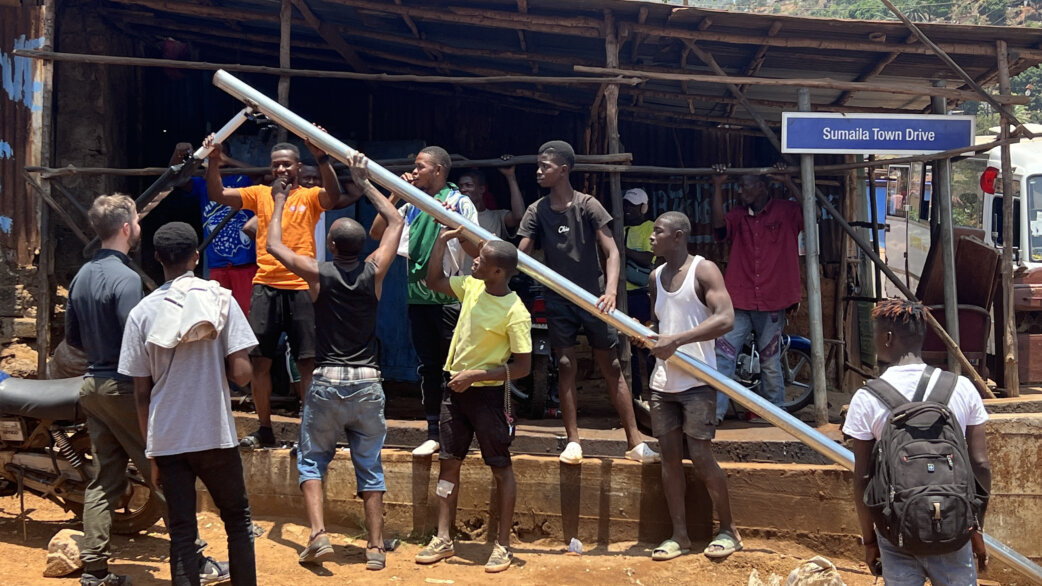 Community members in Freetown, Sierra Leone lifting and installing a solar-powered LED streetlight pole, with a street sign reading "Sumaila Town Drive" visible in the background.