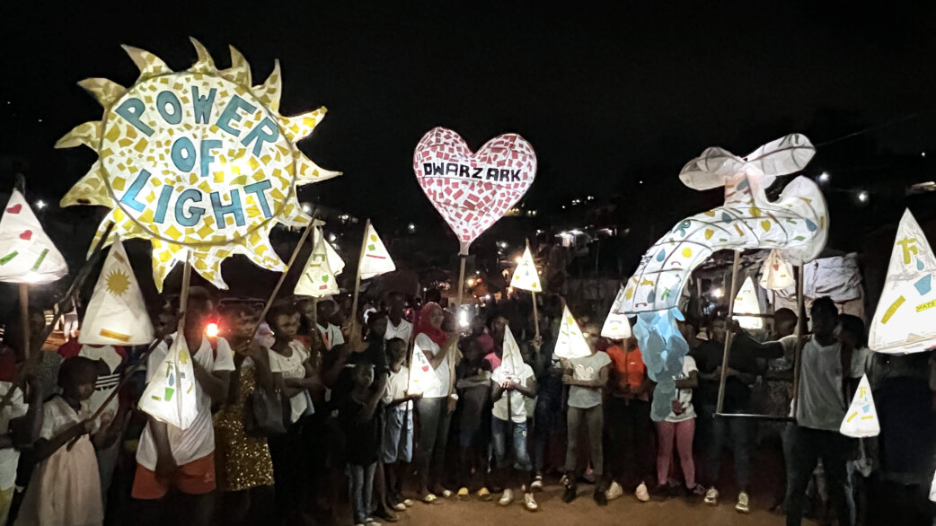 A nighttime lantern parade in Freetown, Sierra Leone, featuring illuminated paper lanterns including a sun-shaped design reading "Power of Light" and a heart reading "Dwarzark", held by community members.