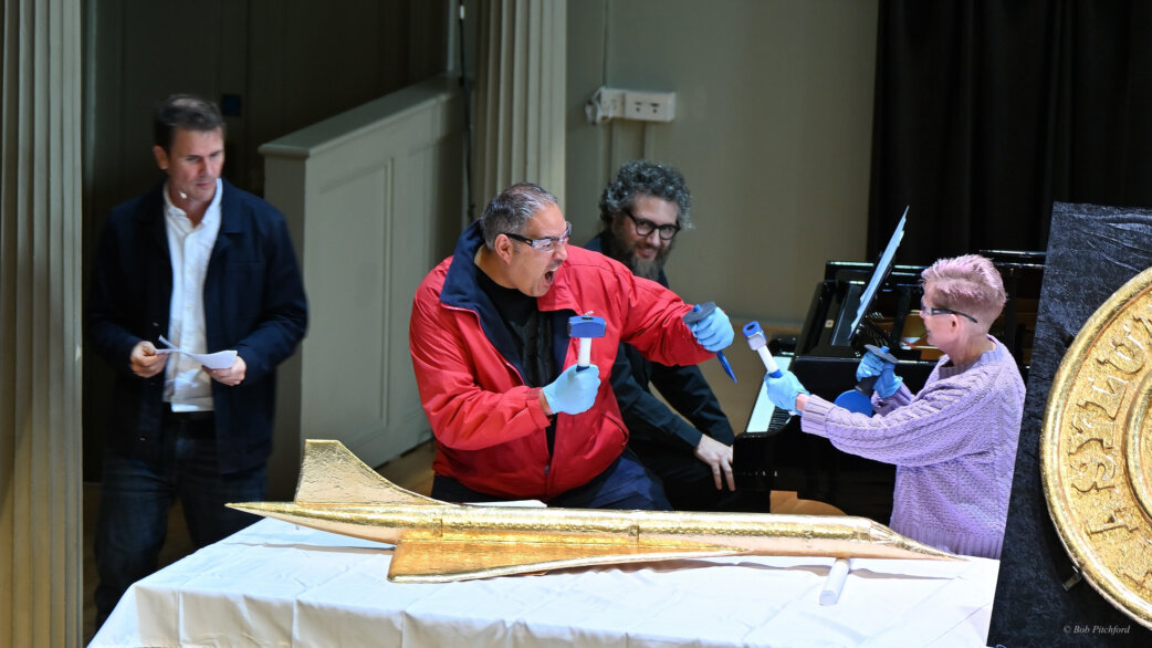 A man in a red jacket uses a chisel to break apart a gold-foil-wrapped chocolate Concorde replica, approximately 1.5 metres long, while a child assists and others watch nearby.