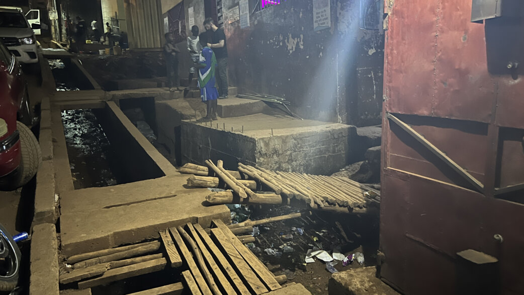 A nighttime street scene in Freetown showing an open drainage channel with makeshift wooden plank crossings, people standing nearby, and informal settlement buildings illuminated by streetlighting.