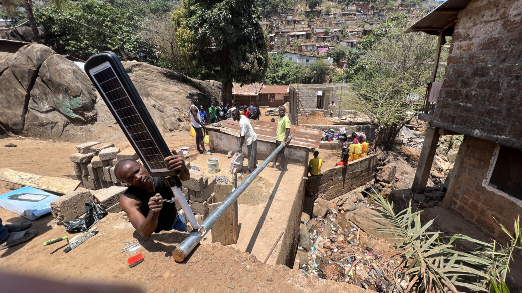 A man installs a solar-powered LED streetlight on a metal pole in an informal settlement in Freetown, Sierra Leone, with densely packed hillside housing visible in the background.