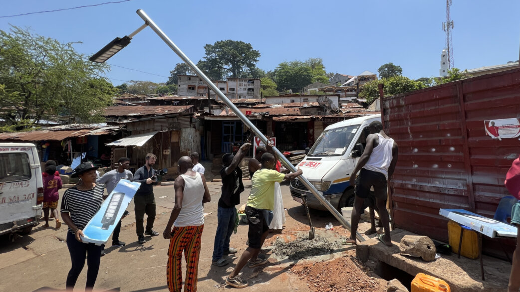 Community members work together to raise a solar-powered LED streetlight pole in an informal settlement in Freetown, Sierra Leone, with corrugated iron buildings visible behind.