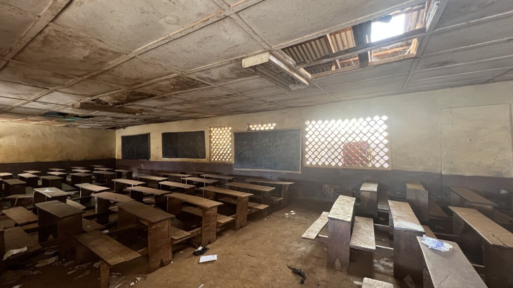 A dimly lit classroom in Freetown, Sierra Leone, with rows of wooden desks, chalkboards on the walls, and damaged ceiling panels allowing light through gaps.