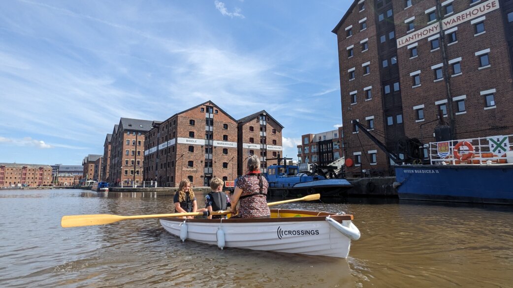 Three people row a white wooden boat named Crossings through brown water at Gloucester Docks, passing historic brick warehouses including Llanthony Warehouse, on a sunny day.
