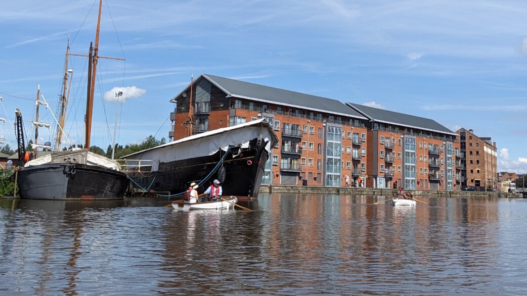 Two people in a white rowing boat on a waterway, part of the Crossings installation, with historic sailing vessels and modern red brick apartments in the background at Gloucester docks.