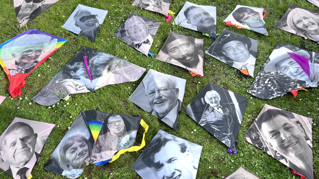 Black and white portrait photographs printed on kites laid out on grass, showing faces of people who died during Covid-19, with colorful kite tails visible.