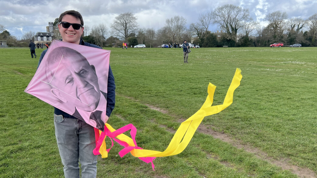 A smiling man in sunglasses holds a pink kite featuring a black-and-white portrait of an elderly person, with yellow and pink ribbon tails, on a grassy field.