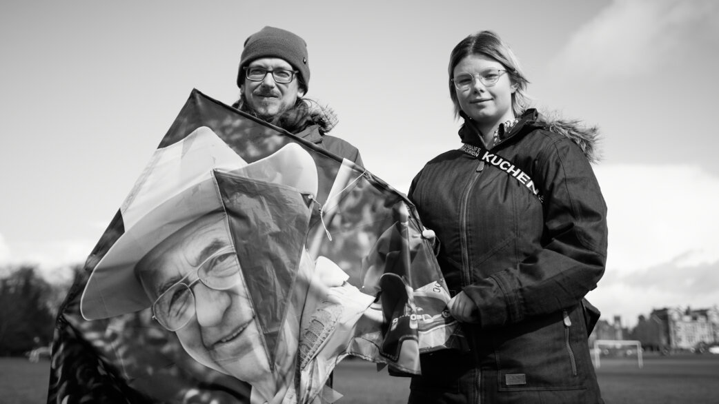 Black and white photograph of two people standing outdoors holding a kite printed with an elderly person's portrait, part of a memorial event on Bristol Downs.