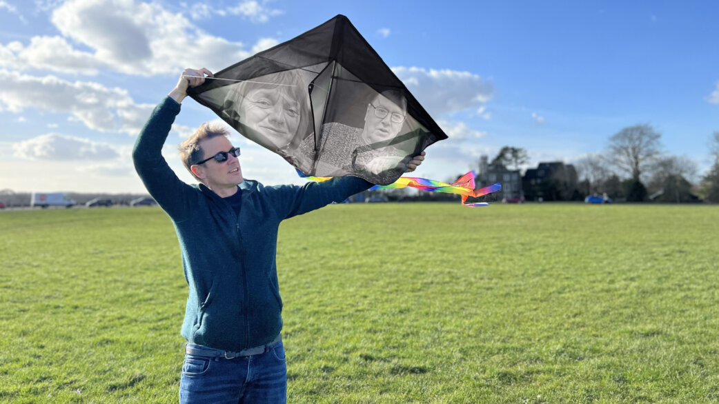 A man in a blue jumper holds up a black diamond-shaped kite printed with two portrait photographs, with a rainbow tail, on a grassy park on a sunny day.