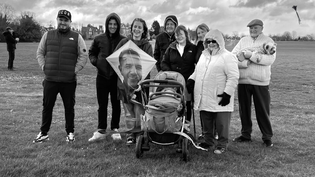 A group of nine people stand on a grassy field holding a diamond-shaped kite printed with a man's smiling face, with a baby stroller and small dog visible.