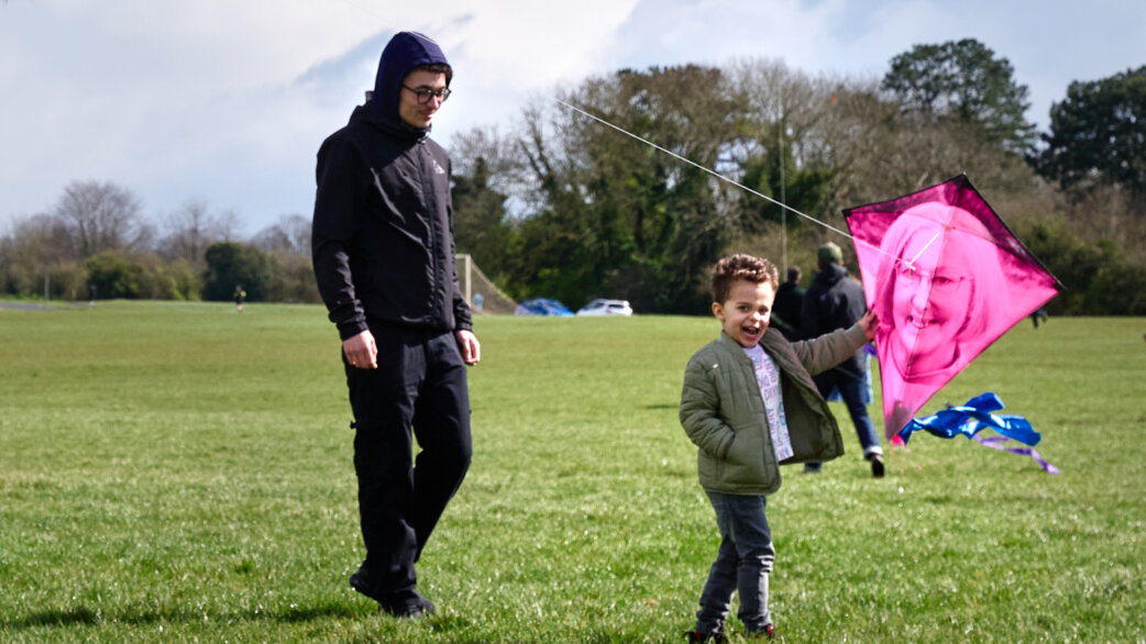 A young boy joyfully holds a pink kite printed with a woman's face while an adult in a dark hoodie walks beside him on a grassy field during the Ascension memorial event.