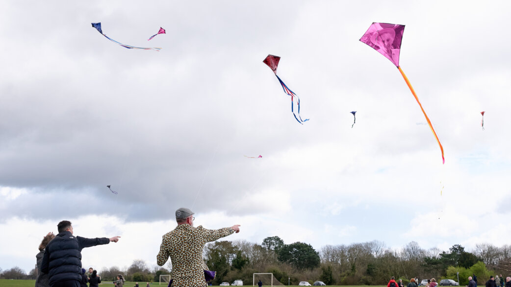 People fly colorful diamond-shaped kites on a grassy field under overcast skies. One pink kite displays a photograph of a person. Part of a Covid-19 memorial event in Bristol.