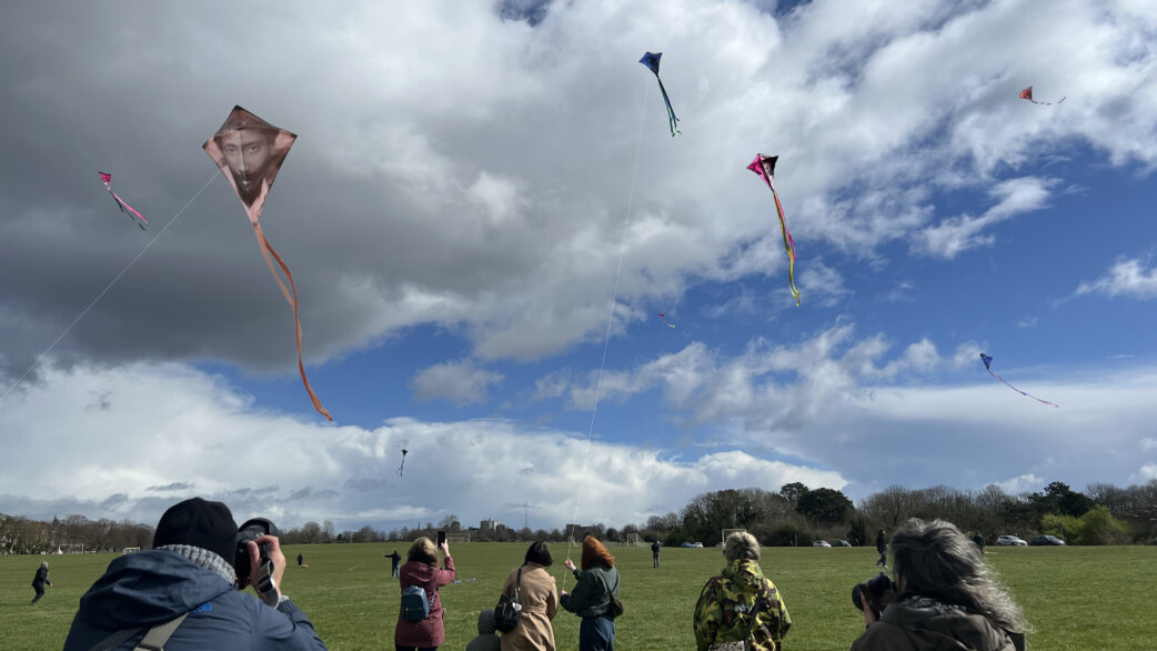 People on a grassy field fly colorful kites against a dramatic cloudy sky. One prominent diamond-shaped kite displays a portrait of a person, with long orange streamers trailing below.