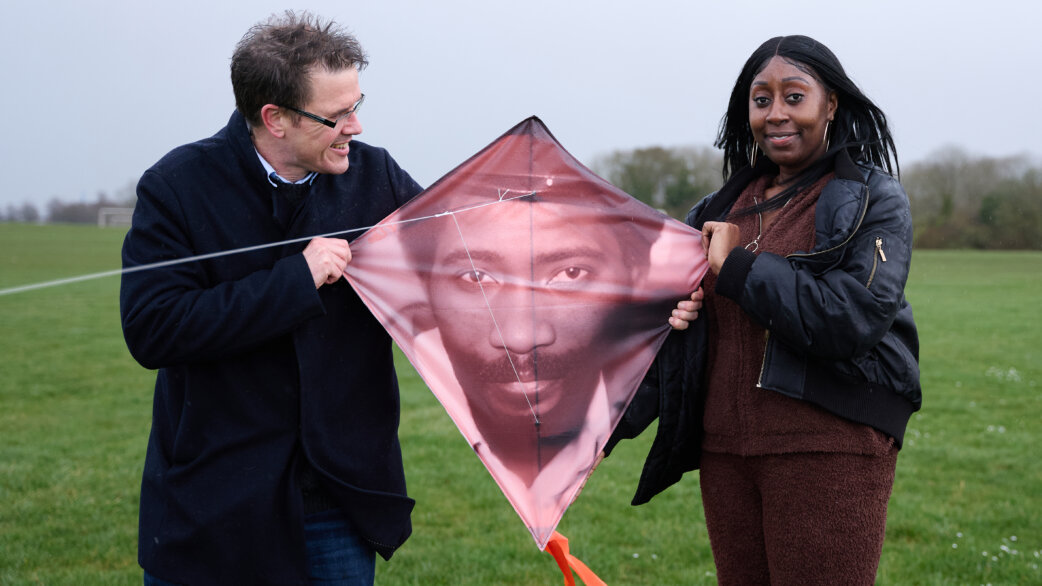 Two people hold a diamond-shaped kite featuring a printed portrait of a man's face in muted pink tones, standing on a green field on an overcast day.