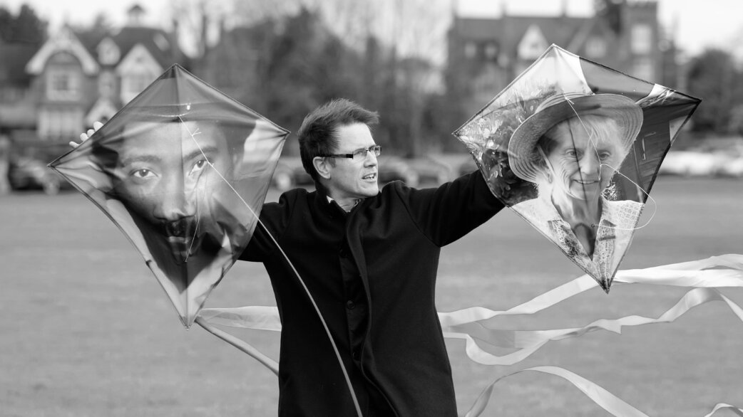 A man in a dark coat holds two diamond-shaped kites with printed portraits of people, standing in a grassy field with streamers flowing below. Black and white photograph.