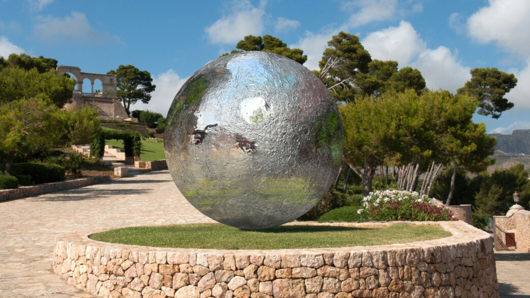 A large reflective stainless steel sphere resembling the Moon, with detailed crater textures, displayed on a grass mound with stone edging in a Mediterranean garden setting.