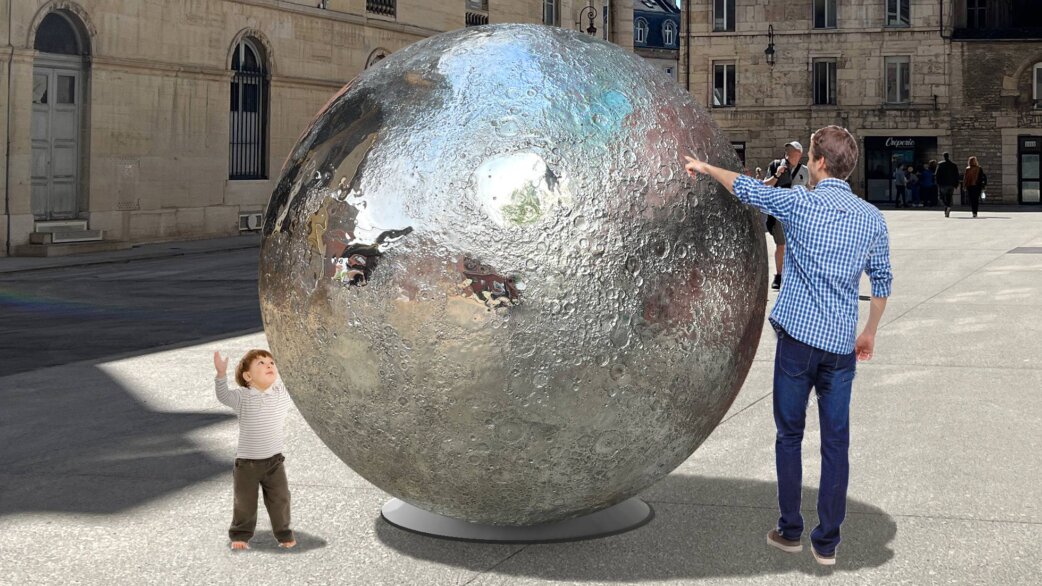 A large reflective stainless steel sphere with detailed lunar surface craters sits in a European plaza. A toddler reaches up toward it while an adult points at its surface.