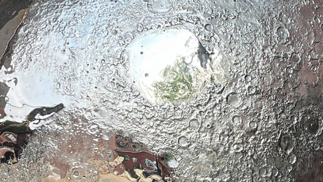 A reflective stainless steel sculpture of the Moon showing detailed crater textures, with surrounding exhibition space visible in its mirrored surface.