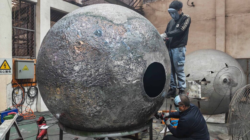 Two workers in a factory workshop polish and finish a large spherical stainless steel moon sculpture, approximately 2 meters in diameter, with detailed crater textures covering its surface.