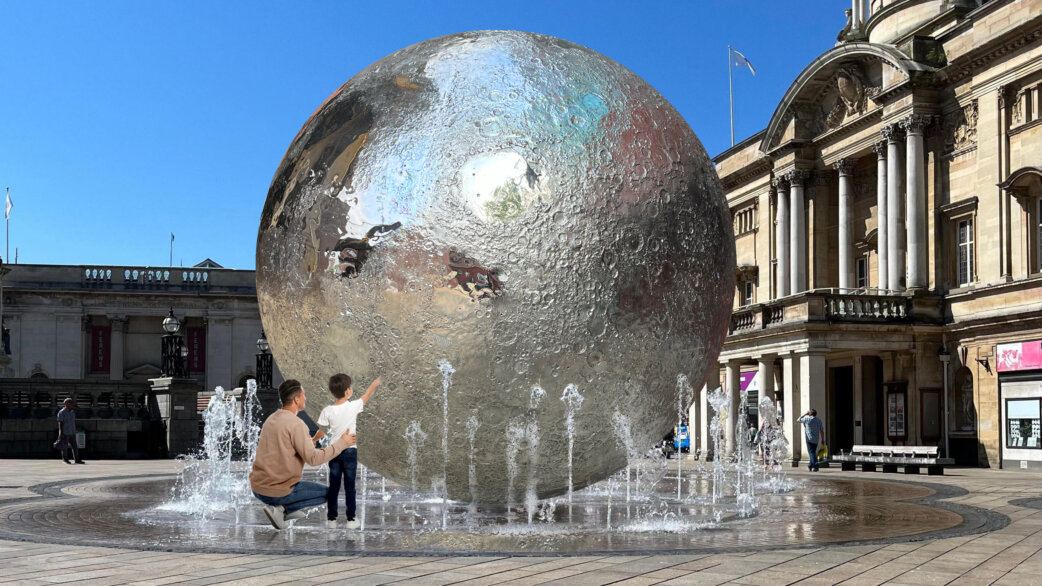 A large reflective stainless steel moon sculpture with detailed crater textures sits in a public square fountain, with an adult and child reaching toward it. Classical buildings visible behind.