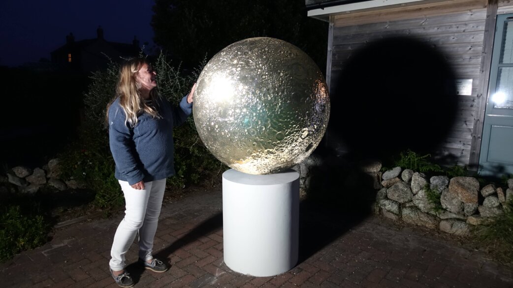 A woman touches a large reflective stainless steel moon sculpture on a white pedestal at night, its cratered surface gleaming under artificial light at an outdoor display.