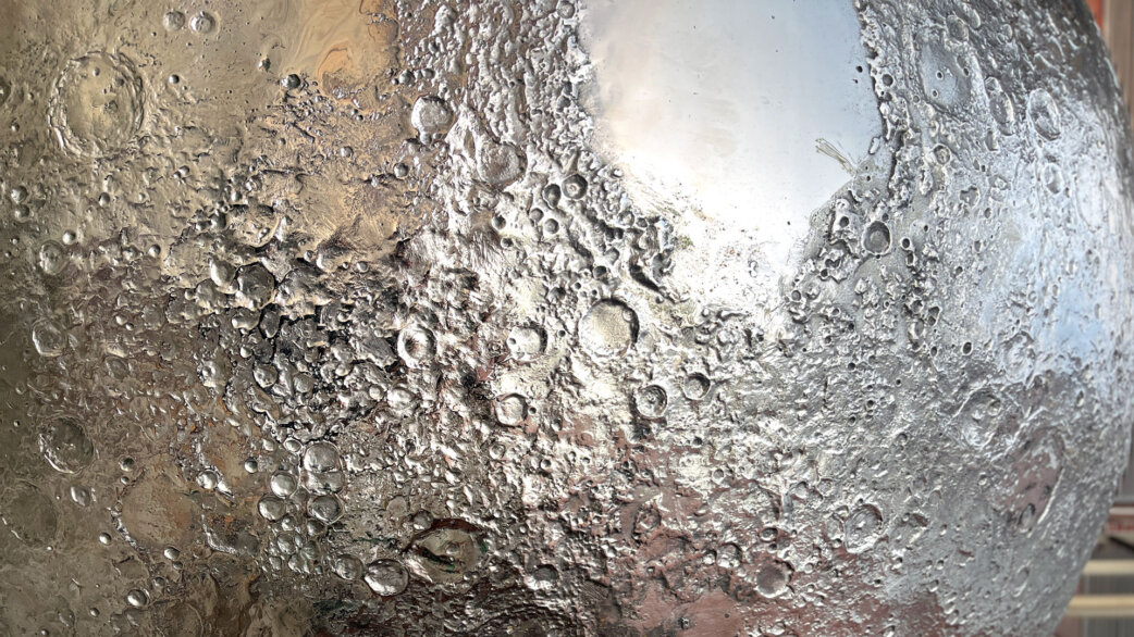 A detailed stainless steel sculpture of the Moon's surface showing craters, valleys, and mountains, with reflective mirror finish catching surrounding light and environment.