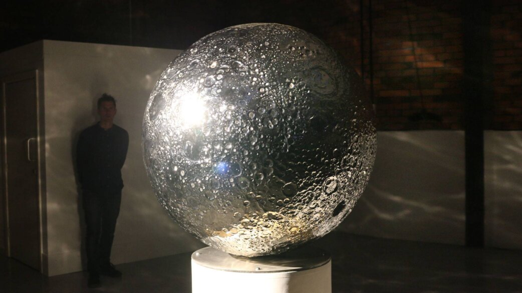 A reflective stainless steel sphere approximately 1 meter in diameter depicting the Moon's cratered surface, mounted on a white pedestal in a dimly lit gallery space with a person standing nearby.