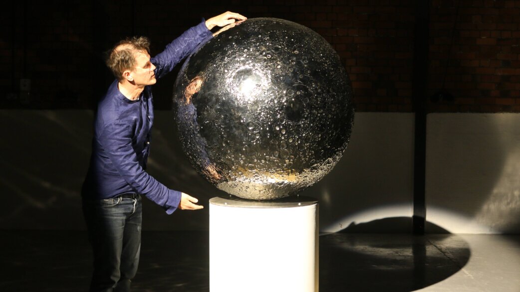 A man in a blue shirt touches a reflective stainless steel sphere approximately one metre in diameter, depicting the Moon's cratered surface, displayed on a white pedestal.