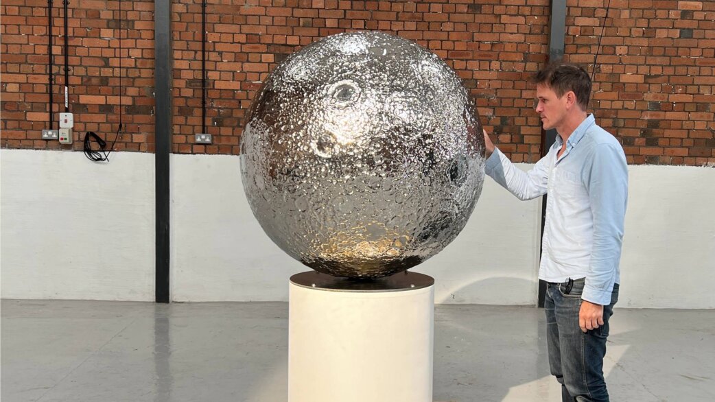 A man in a light blue shirt touches a reflective stainless steel sphere approximately one metre in diameter, featuring detailed lunar surface craters, displayed on a white cylindrical plinth.
