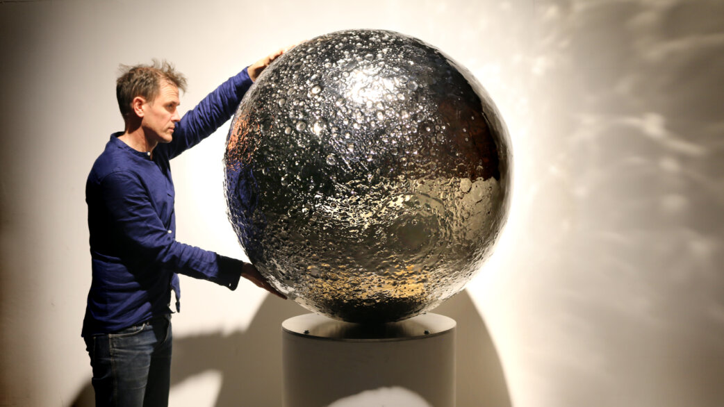 A man in a blue shirt touches a reflective stainless steel sculpture of the Moon, approximately 1 meter in diameter, mounted on a cylindrical pedestal. The polished surface shows detailed craters and topography.