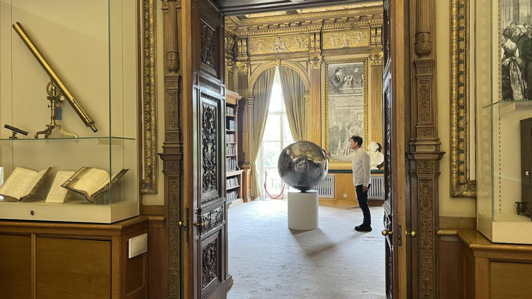 A visitor examines a large reflective stainless steel Moon sculpture displayed on a white pedestal in an ornate historic library room with gilded frames, antique telescopes, and old books.