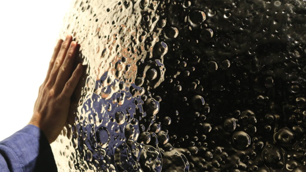 A hand touches a reflective stainless steel sculpture of the Moon, approximately 1 meter in diameter, showing detailed craters and surface topology based on NASA data.