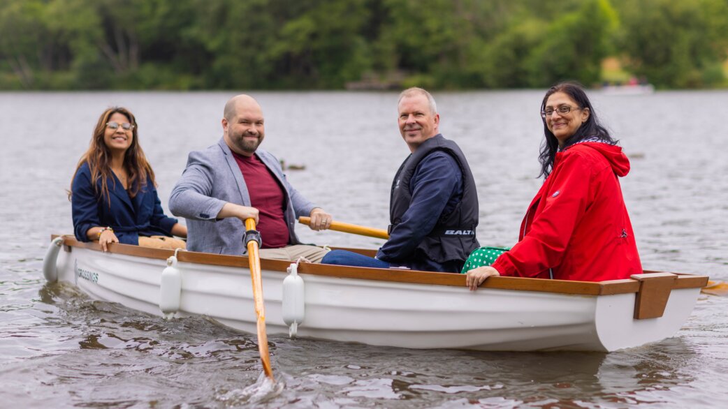 Four people smiling while rowing a white wooden boat on a lake, part of Luke Jerram's Crossings installation where boats play audio stories to passengers.
