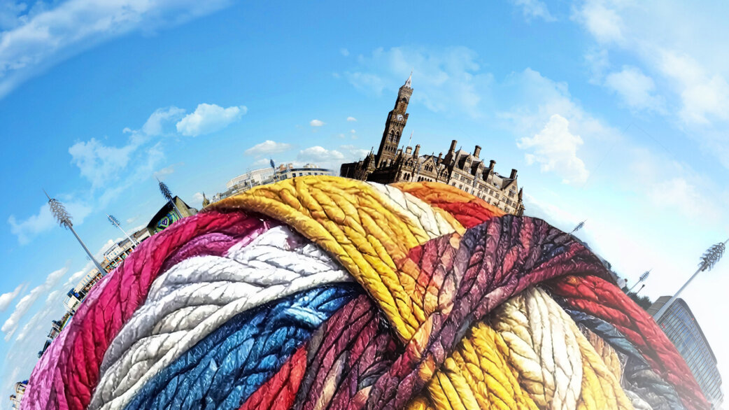 A colourful three-metre yarn ball made from plaited fabric in red, yellow, blue and cream, photographed with Bradford City Hall visible behind against a blue sky. A fisheye lens is used so it looks like the yarn ball is the ground with the buildings resting on top of it.