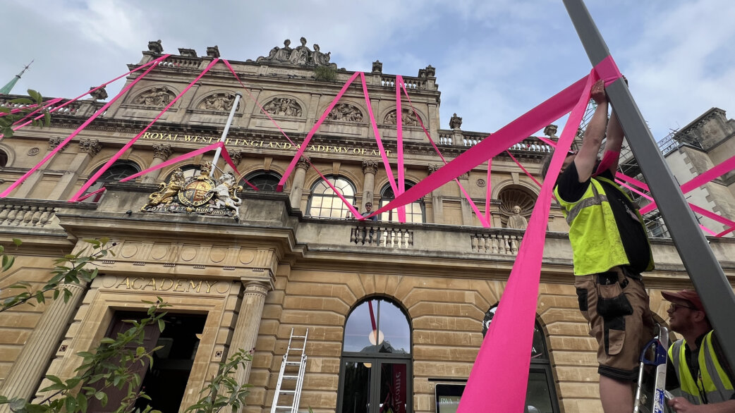 Workers in high-visibility vests attach bright magenta ribbon to a lamppost outside the Royal West of England Academy, with ribbons stretching across the classical stone façade above.