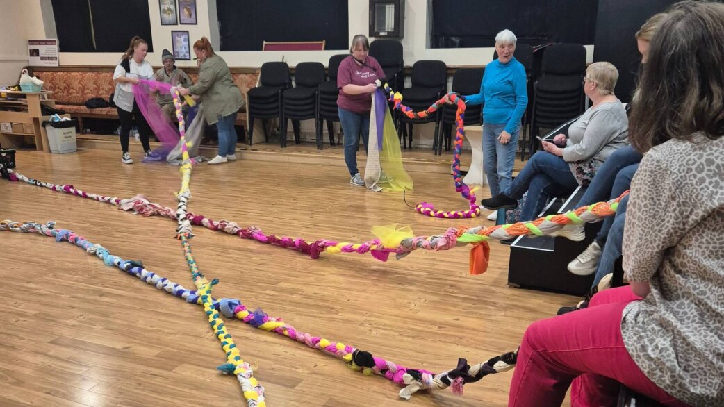 A community workshop in a hall where participants hold and display long colourful plaited ropes made from fabric, laid out across wooden flooring.
