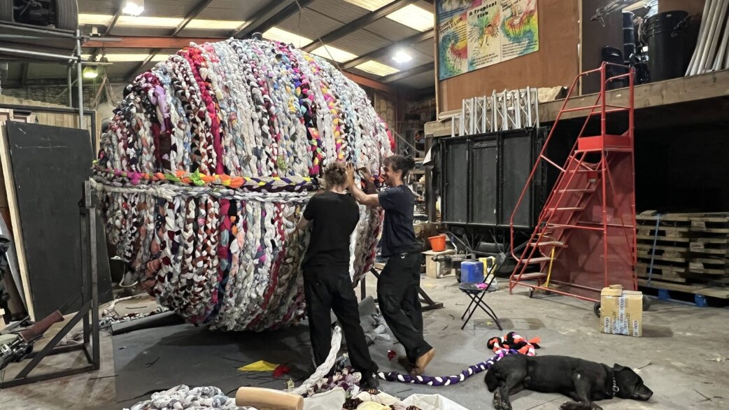Two people work on a large spherical sculpture, approximately three metres tall, made from colourful braided fabric ropes in a workshop setting. A black dog rests nearby.