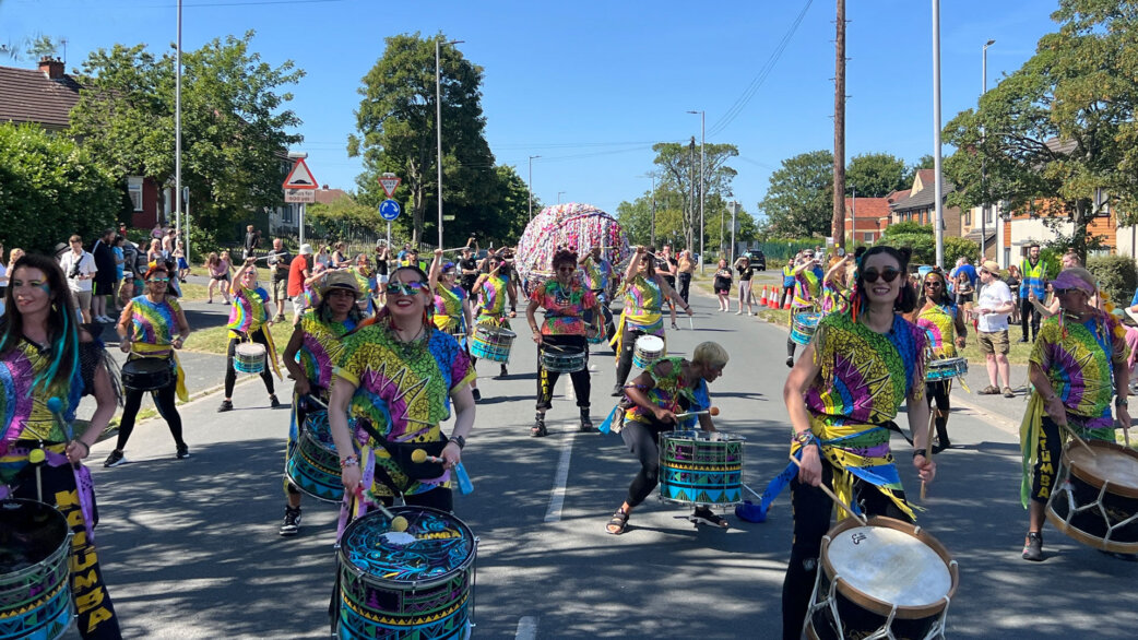 A colourful samba drumming band in vibrant tie-dye costumes parades through a residential street, with a three-metre-wide yarn ball visible behind them on a sunny day.