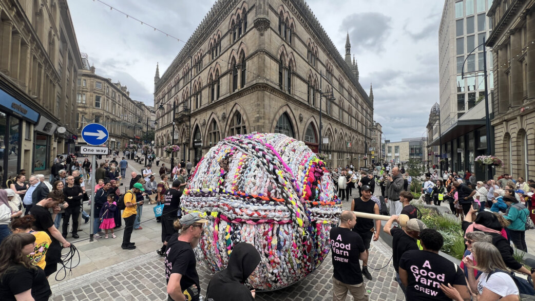 A giant three-metre yarn ball made from colourful plaited fabric being rolled through Bradford city centre by crew members wearing black t-shirts, with Victorian Gothic architecture behind.