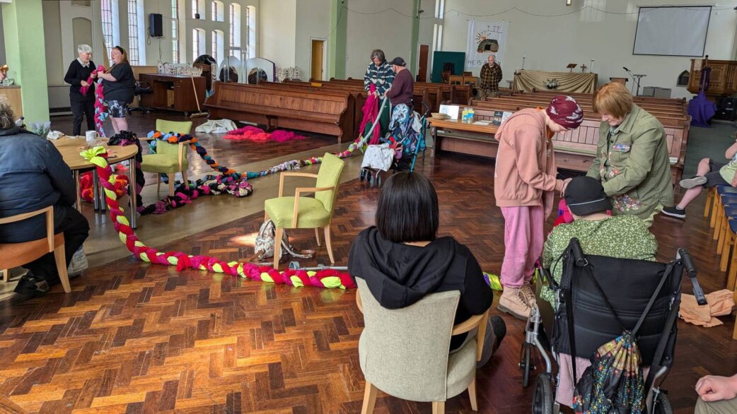 Community members in a church hall creating a colourful plaited rope from donated fabric, with the multicoloured textile snake winding across the wooden floor between chairs.